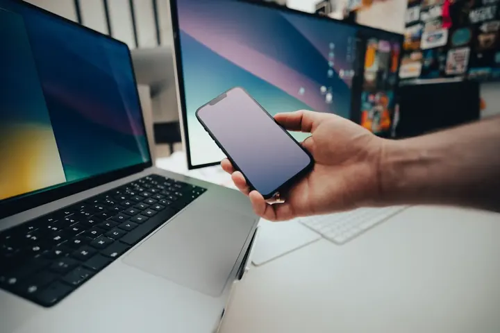 Mão segurando um smartphone diante de um notebook e um monitor